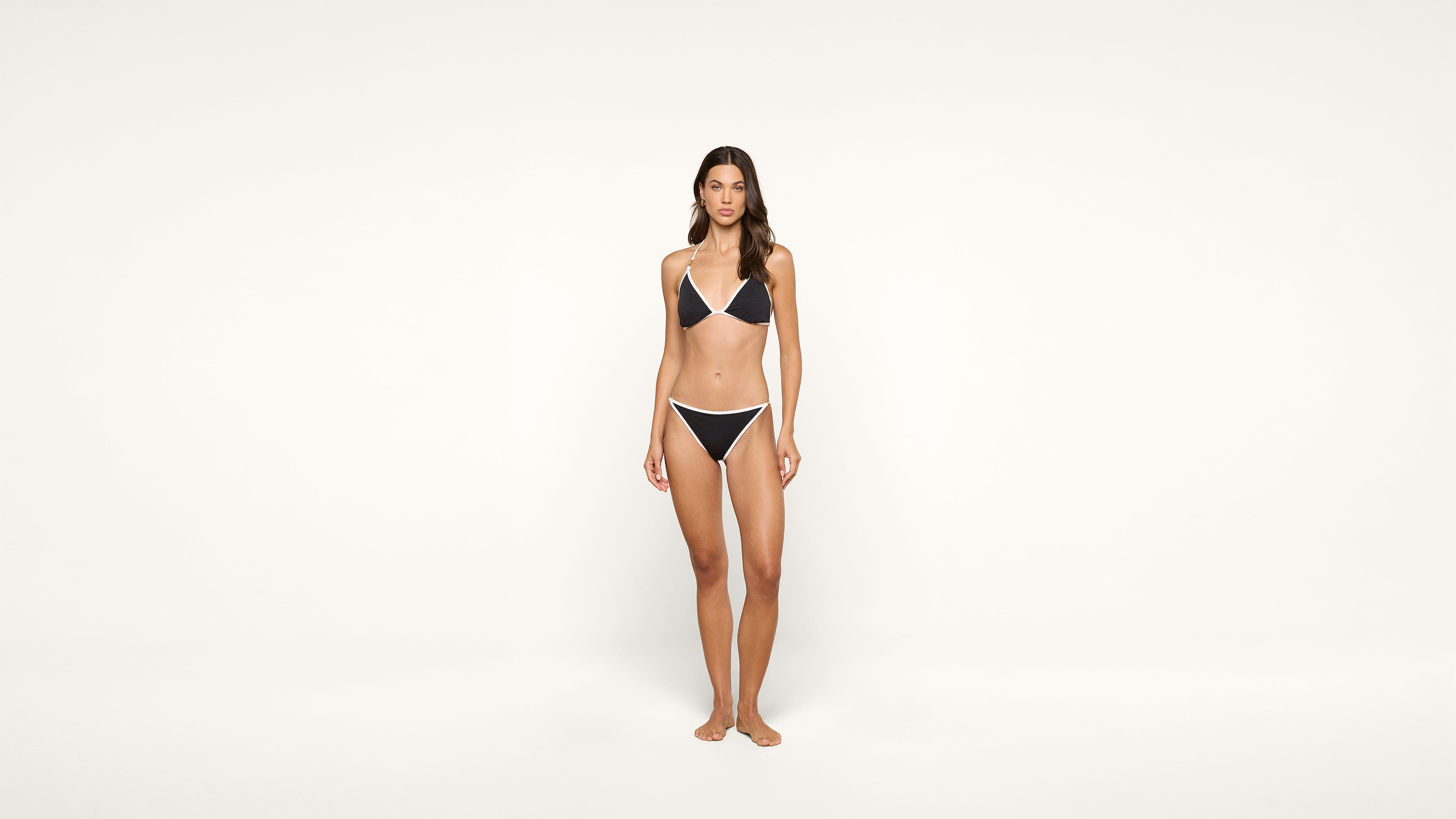 A woman stands barefoot against a plain white background, wearing a black bikini with white trim.