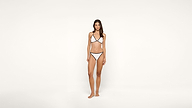 A woman stands barefoot against a plain white background, wearing a white bikini with black trim.