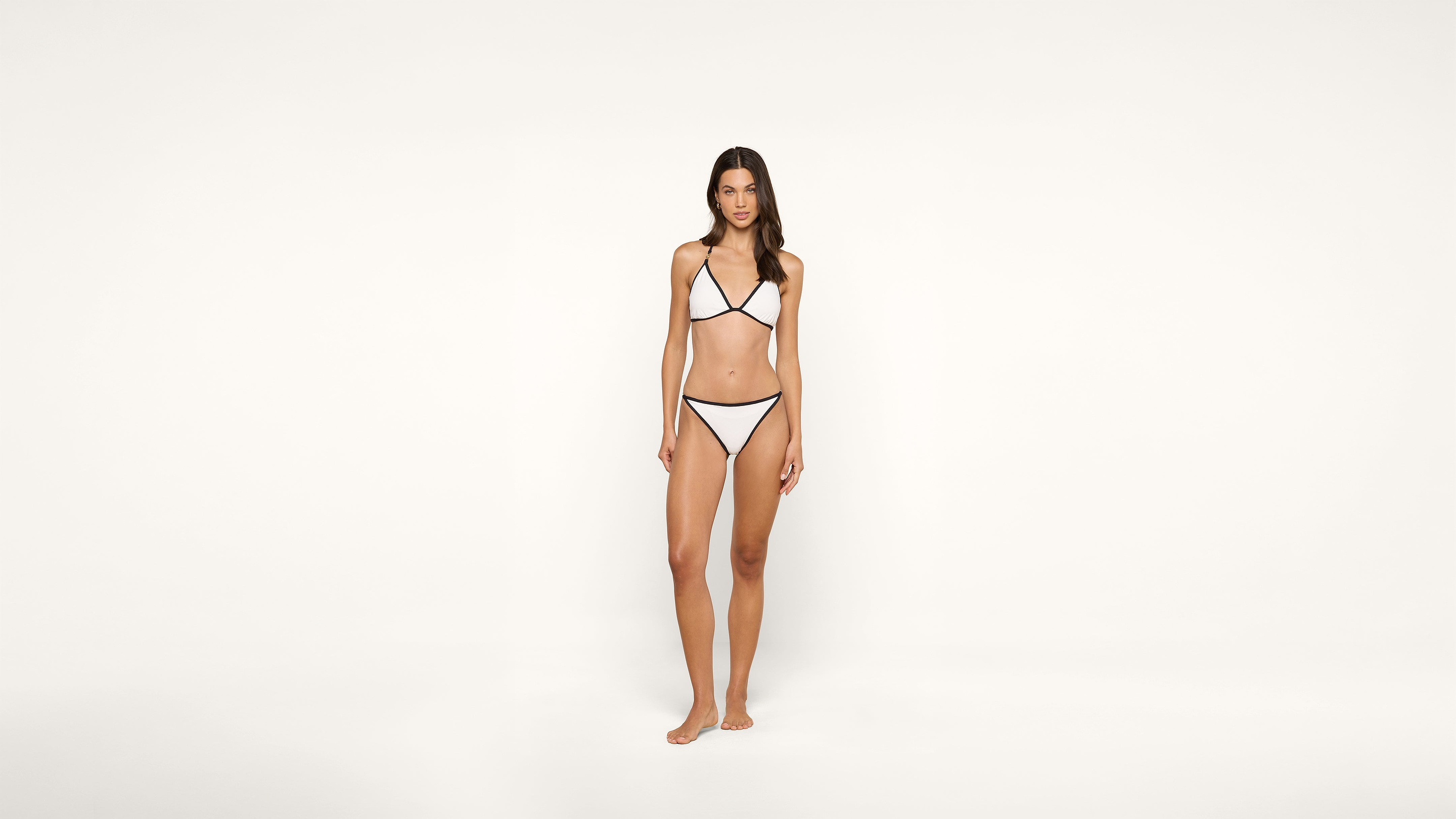 A woman stands barefoot against a plain white background, wearing a white bikini with black trim.