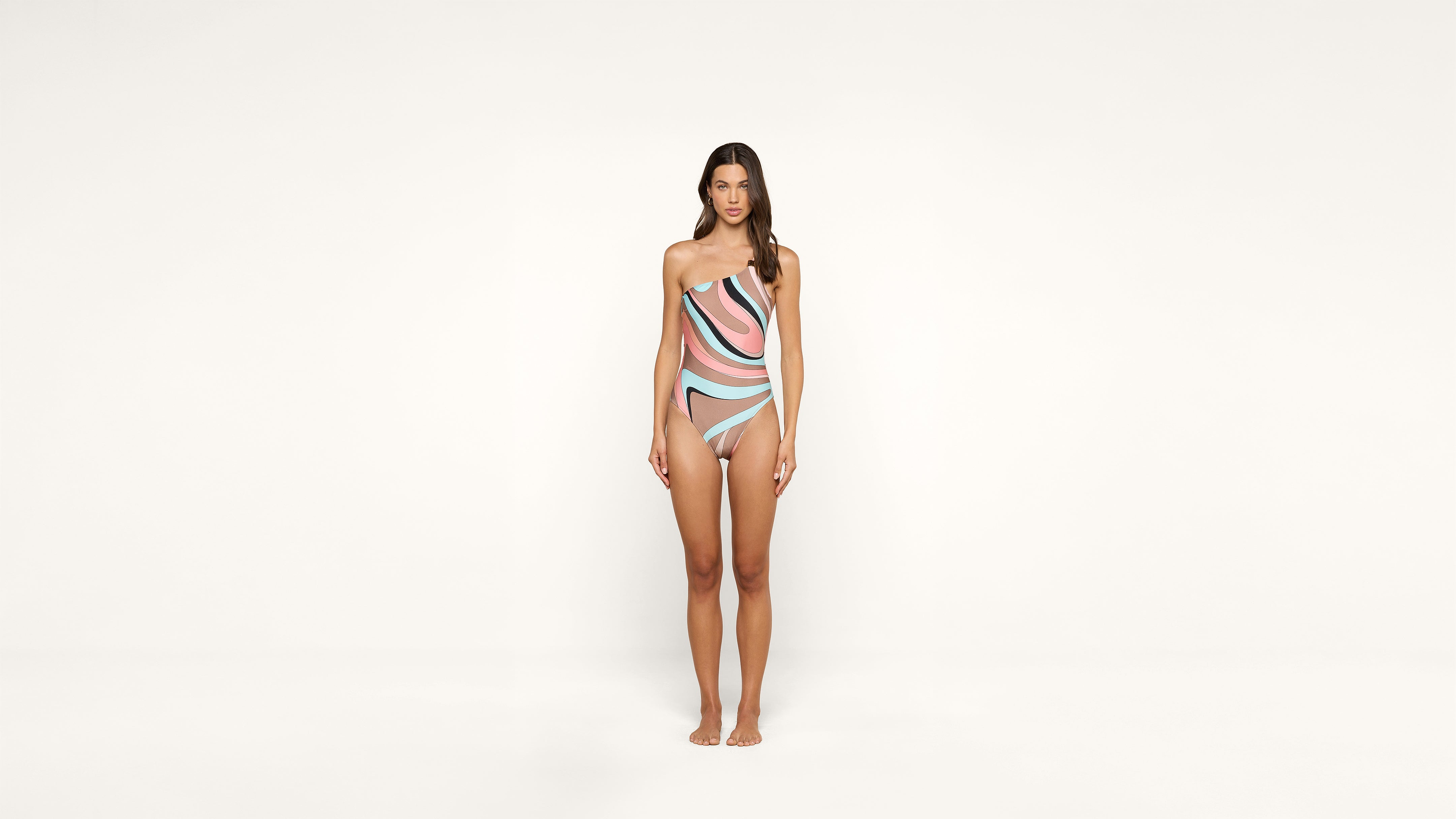 A woman stands barefoot against a plain white background, wearing a one-shoulder swimsuit with pink, black, and light blue wavy patterns. She faces forward with her arms by her sides.