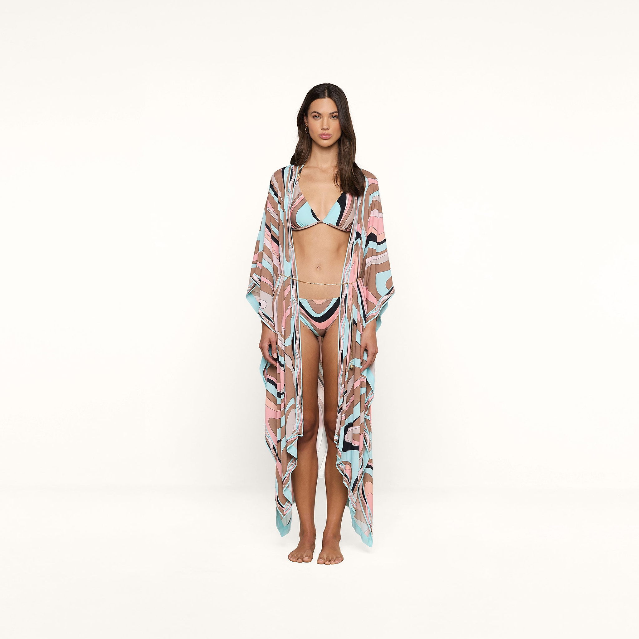 A woman stands barefoot wearing beachwear: a bikini paired with the Kara Kimono Cover-Up, crafted from viscose crepe in pastel blue, pink, black, and white swirls, against a plain white background.