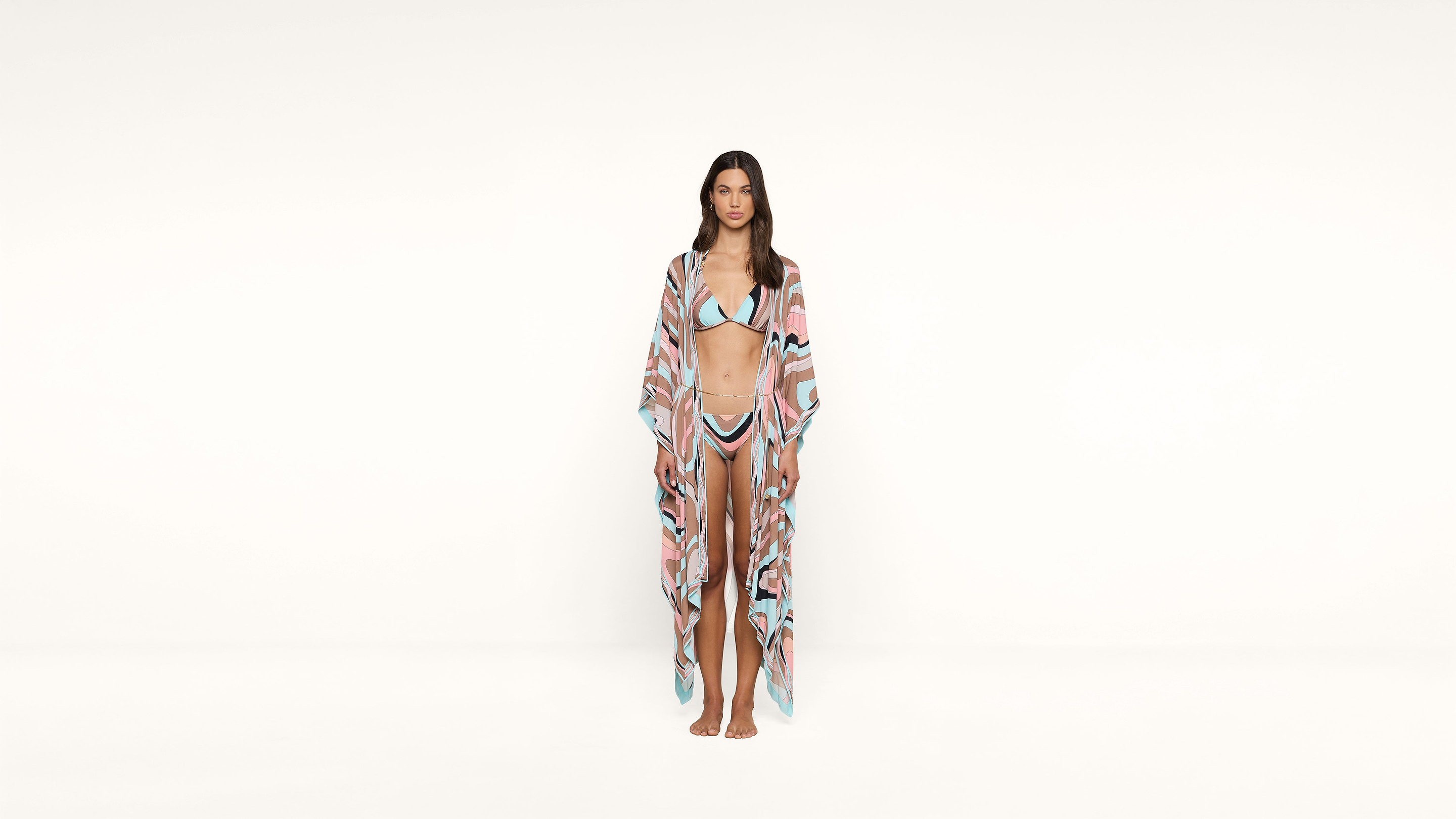A woman stands barefoot against a plain white background, wearing a pastel-colored bikini with a matching long, sheer cover-up.