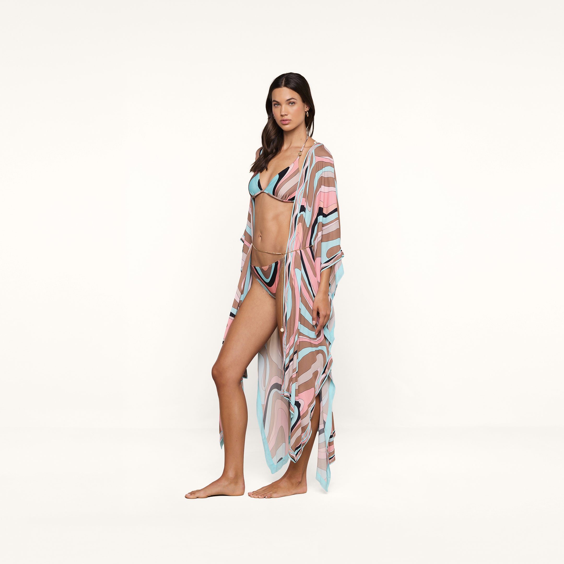 A woman stands barefoot against a plain white background, looking at the camera with a neutral expression, wearing a pastel patterned bikini paired with the Kara Kimono Cover-Up in matching viscose crepe.