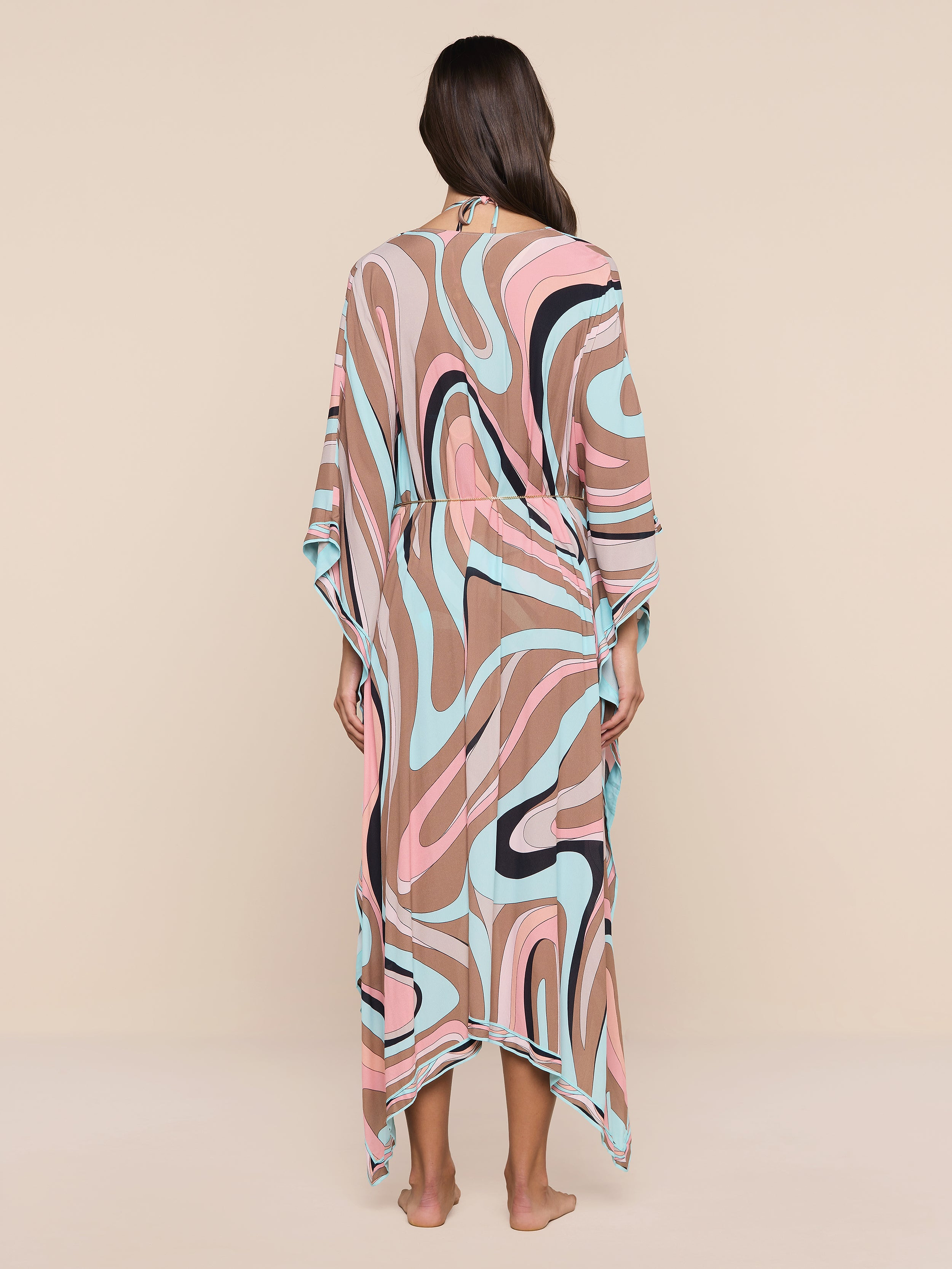 A woman stands barefoot facing away, wearing the Kara Kimono Cover-Up crafted from flowing viscose crepe with a pink, blue, beige, black, and white abstract swirl pattern. The plain white background highlights this stylish piece—perfect as a beachwear cover-up.