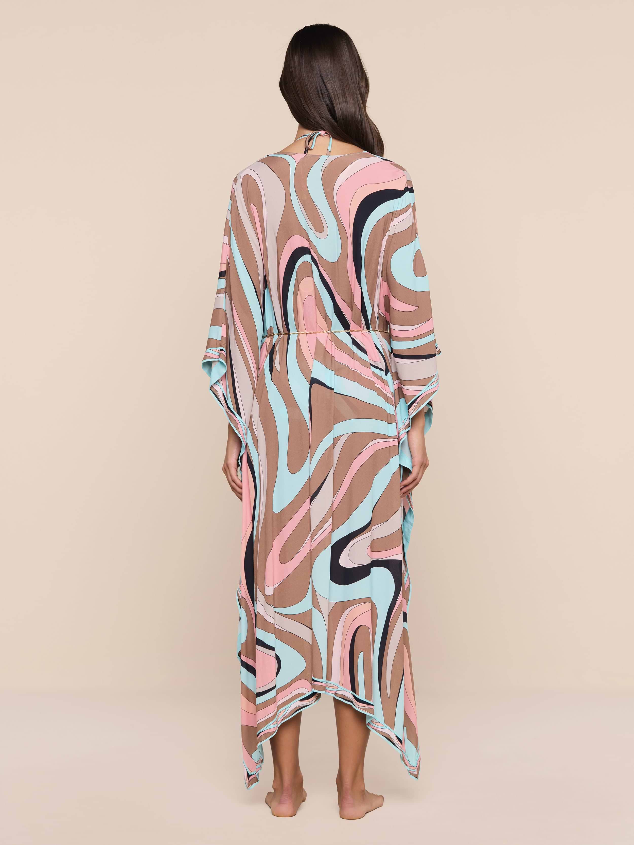 A woman stands barefoot facing away, wearing the Kara Kimono Cover-Up crafted from flowing viscose crepe with a pink, blue, beige, black, and white abstract swirl pattern. The plain white background highlights this stylish piece—perfect as a beachwear cover-up.