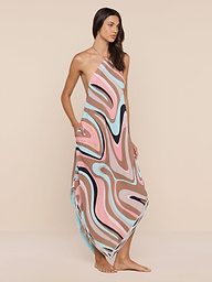 핑크, 브라운, 블루, 블랙 톤의 추상적인 소용돌이 프린트가 돋보이는 물에 안전한 크레이프 소재의 원숄더 원피스인 엘리스 커버업 드레스를 입고 맨발로 서 있는 여성이 평범한 베이지색 배경을 배경으로 서 있습니다.