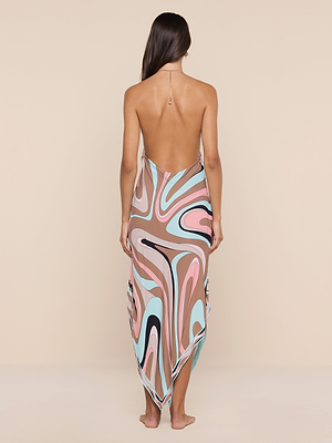 A woman stands barefoot facing away, wearing the Elise Cover-Up Dress—a water-safe crepe halter midi dress featuring a colorful abstract swirl print in pink, brown, blue, black, and white on a beige background.