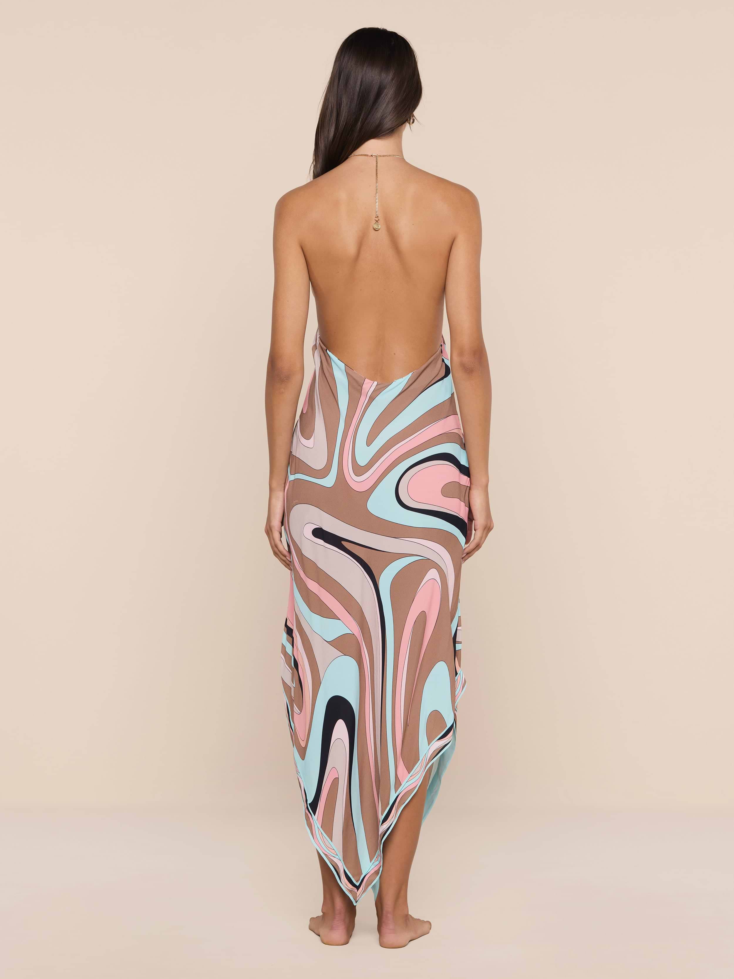베이지색 바탕에 핑크, 브라운, 블루, 블랙, 화이트의 화려한 추상 소용돌이 프린트가 돋보이는 방수 크레이프 홀터 미디 드레스인 엘리스 커버업 드레스를 입은 한 여성이 맨발로 정면을 바라보고 서 있습니다.