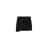 The Chandra Cover-Up Skirt is a black mini with an asymmetrical hem and side draping, accented by a decorative gold hardware buckle on the waistband. Displayed against a plain white background.
