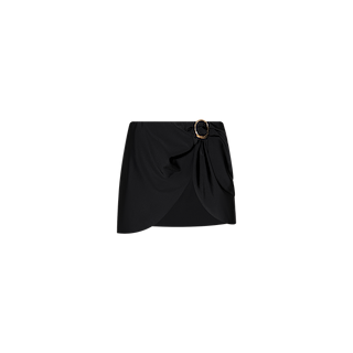 The Chandra Cover-Up Skirt is a black mini skirt with an asymmetrical hem and stylish draped detail, accented by a decorative circular buckle with gold hardware on one side. Shown on a plain white background, it’s the perfect piece to add poolside glam to your look.