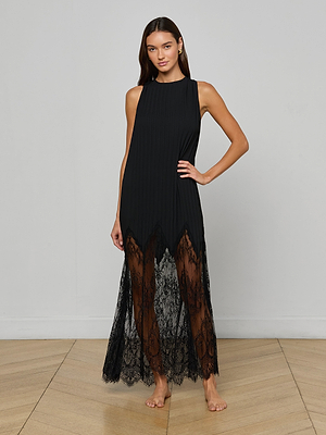 A woman stands barefoot on a wooden floor, wearing the Adalynn Cover-Up Dress—a sleeveless black chiffon piece with a sheer lace skirt and jagged hem—set against a plain light gray wall.