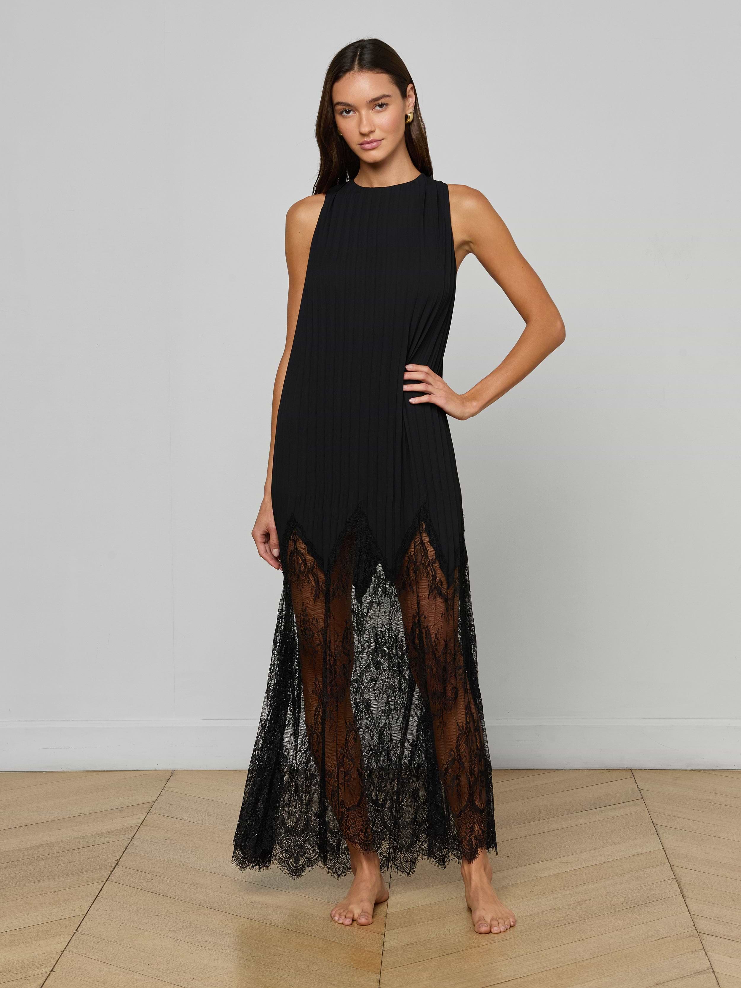 A woman stands barefoot against a plain light background, posing with one hand on her hip and wearing the Adalynn Cover-Up Dress, a sleeveless black lace style featuring sheer lace details at the hem.