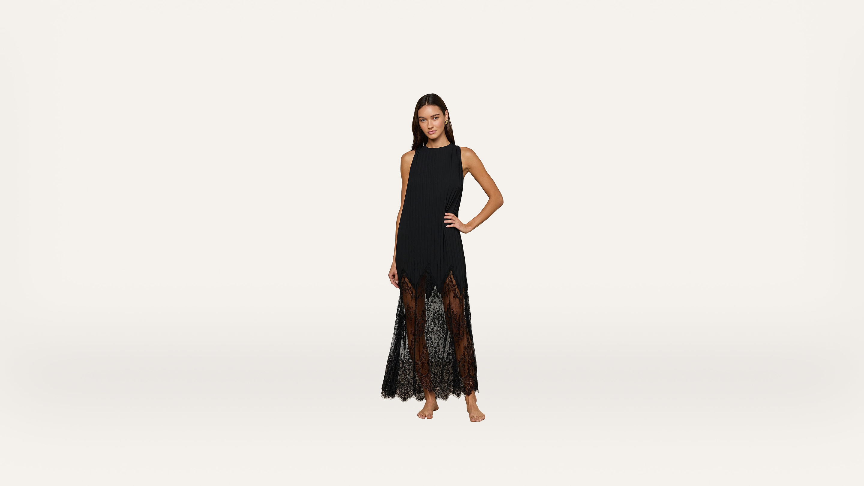 Woman stands barefoot in a sleeveless black dress with a sheer lace skirt, posing with one hand on her hip against a plain white background.
