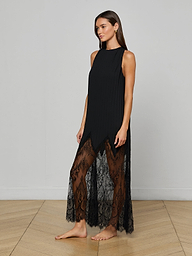 A woman stands barefoot against a plain, light background wearing the Adalynn Cover-Up Dress—an ankle-length, sleeveless black lace dress featuring sheer detailing at the hem.