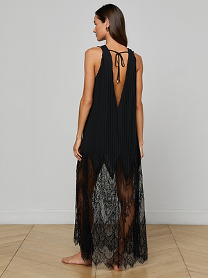 A woman stands barefoot on a wooden floor, facing away with her head turned slightly, wearing the Adalynn Cover-Up Dress—a sleeveless black chiffon piece with a deep V-back, pleated top, and long sheer lace skirt—ideal for chic resort getaways.