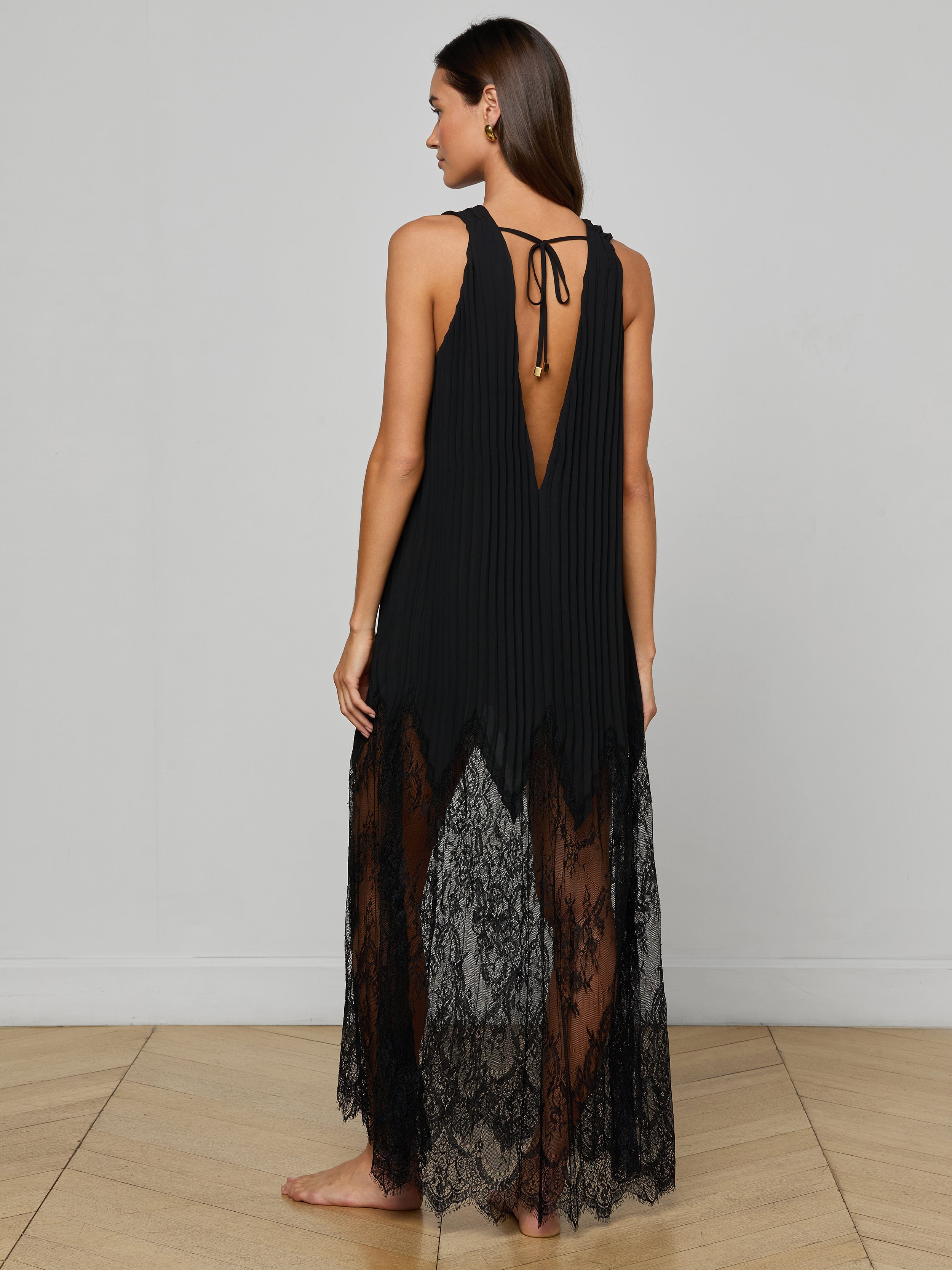A woman stands barefoot and facing away on a plain light background, wearing the Adalynn Cover-Up Dress—a sleeveless black lace dress featuring a deep V-back, pleated upper section, chiffon lower skirt, and a tie detail at the neck.