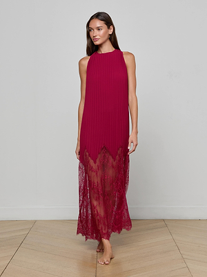 A woman stands barefoot on a wooden floor in the L’AGENCE Adalynn Cover-Up Dress, a sleeveless, ankle-length dark red chiffon piece with a pleated top and lace hem, embodying effortless resort style against a plain gray wall.