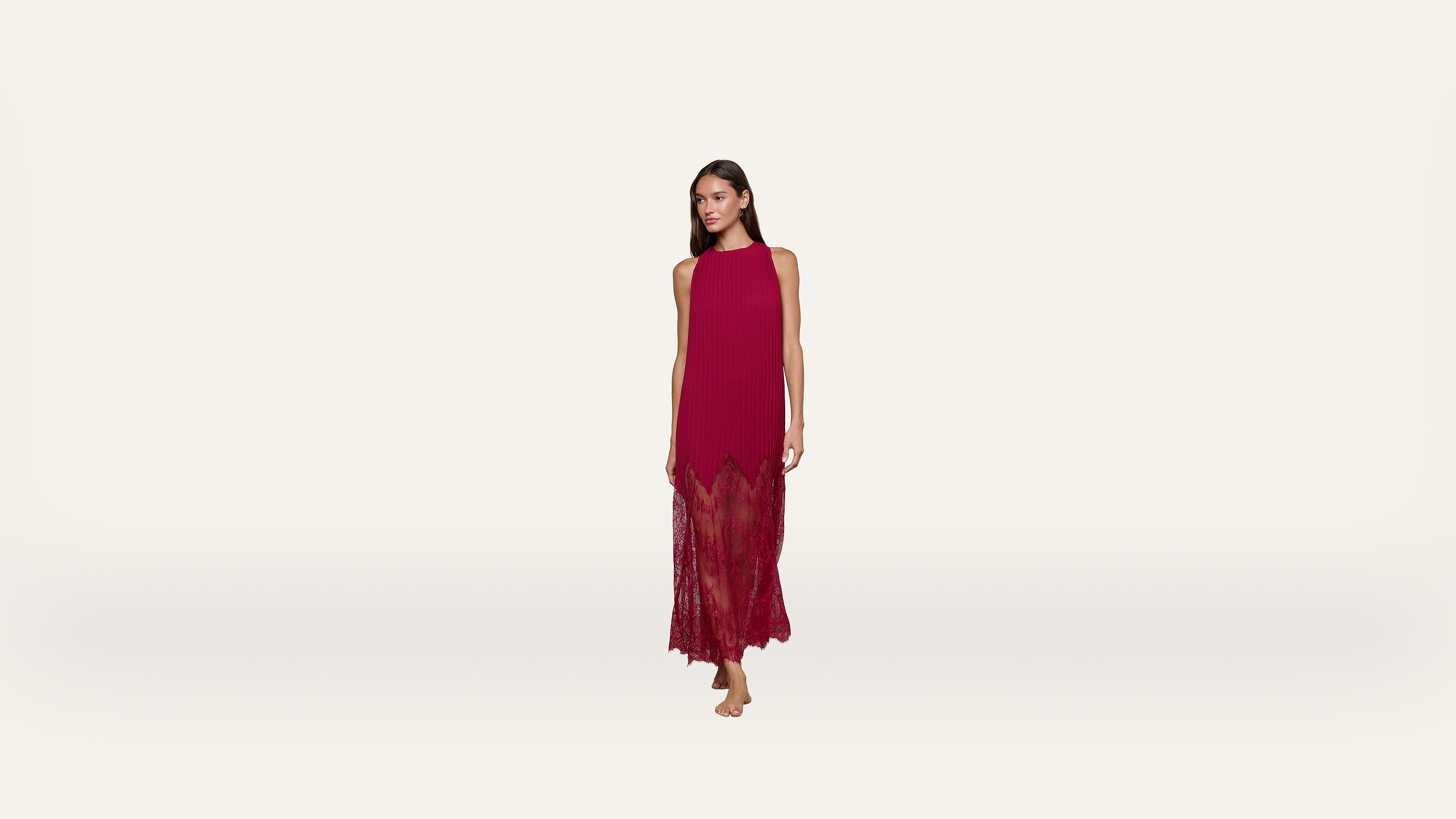 Woman in a sleeveless, floor-length, red dress with lace detailing stands barefoot against a plain white background.