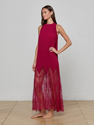 A woman stands barefoot on a wooden floor, wearing the L'AGENCE Adalynn Cover-Up Dress—an ankle-length magenta chiffon piece with a sleeveless pleated top and sheer lace hem. She poses with one hand on her hip against a light gray wall, ideal for resort dressing.