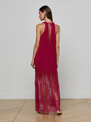 A woman stands on a wooden floor, facing away, wearing the L'AGENCE Adalynn Cover-Up Dress—a sleeveless magenta chiffon dress with pleats, a sheer lace hem, keyhole back with neck tie—against a plain light background.