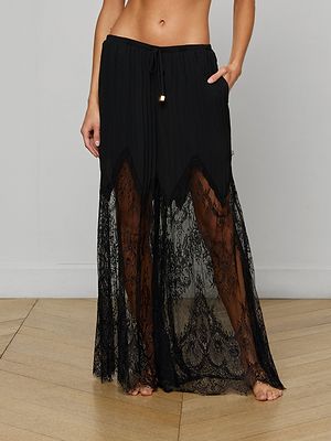 A person stands barefoot on a wooden floor against a plain wall, wearing the Sharon Cover-Up Pant—a long, black, semi-sheer lace pant with pleated chiffon details, a solid waistband, and decorative drawstring.