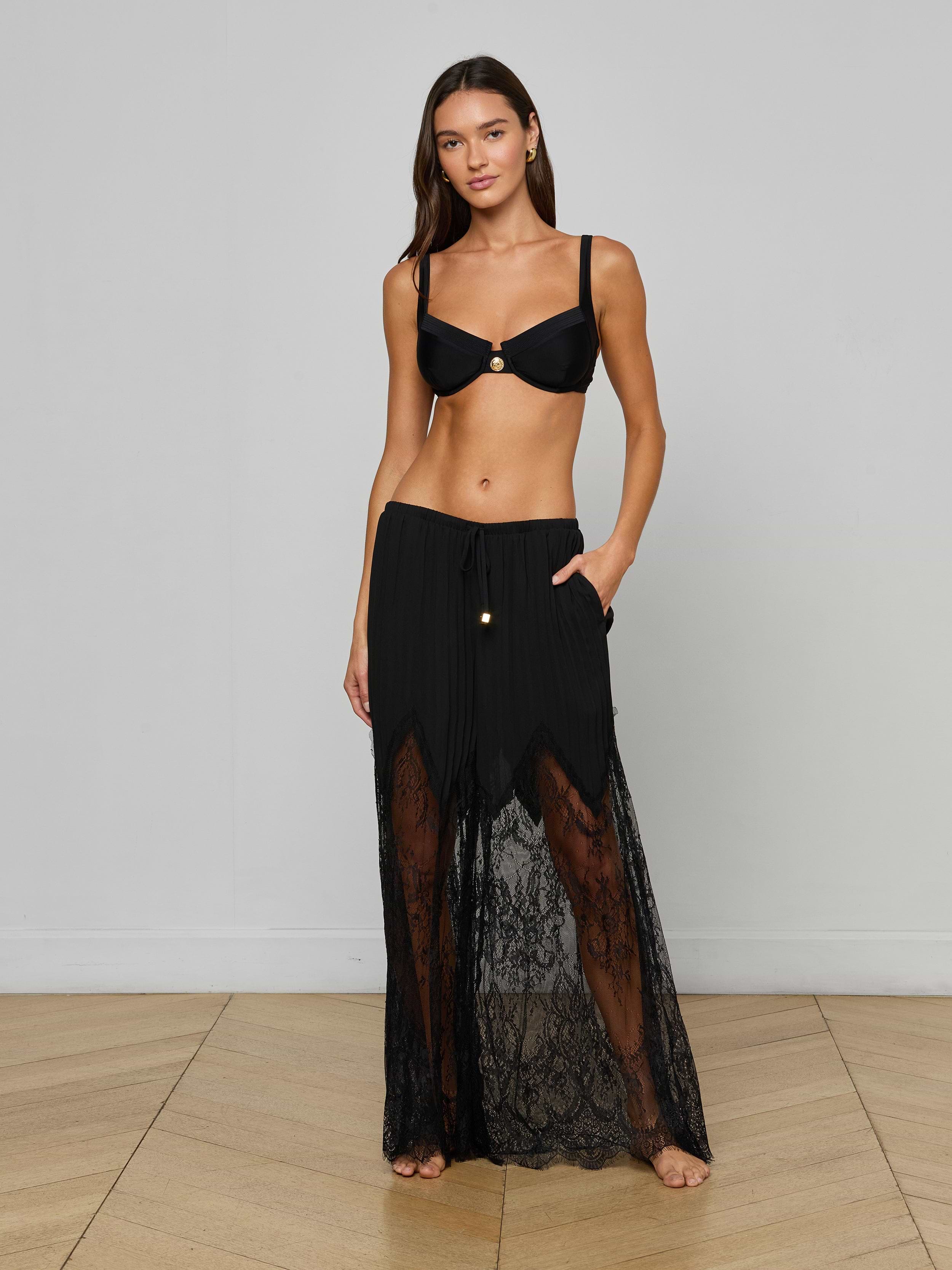 A woman stands barefoot against a plain light background, wearing a black bikini top and the Sharon Cover-Up Pant—a sheer, lacy black wide-leg style—with her left hand in her pocket.