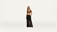 A woman stands barefoot wearing a black bikini top and a sheer black lace maxi skirt with her hands in the skirt pockets against a plain white background.