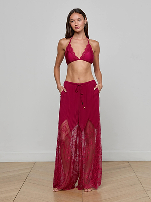 A woman stands indoors exuding effortless elegance in the Sharon Cover-Up Pant—a sheer lace maxi skirt with shorts underneath—paired with a deep red halter bikini top, her long brown hair flowing against a plain light gray wall.