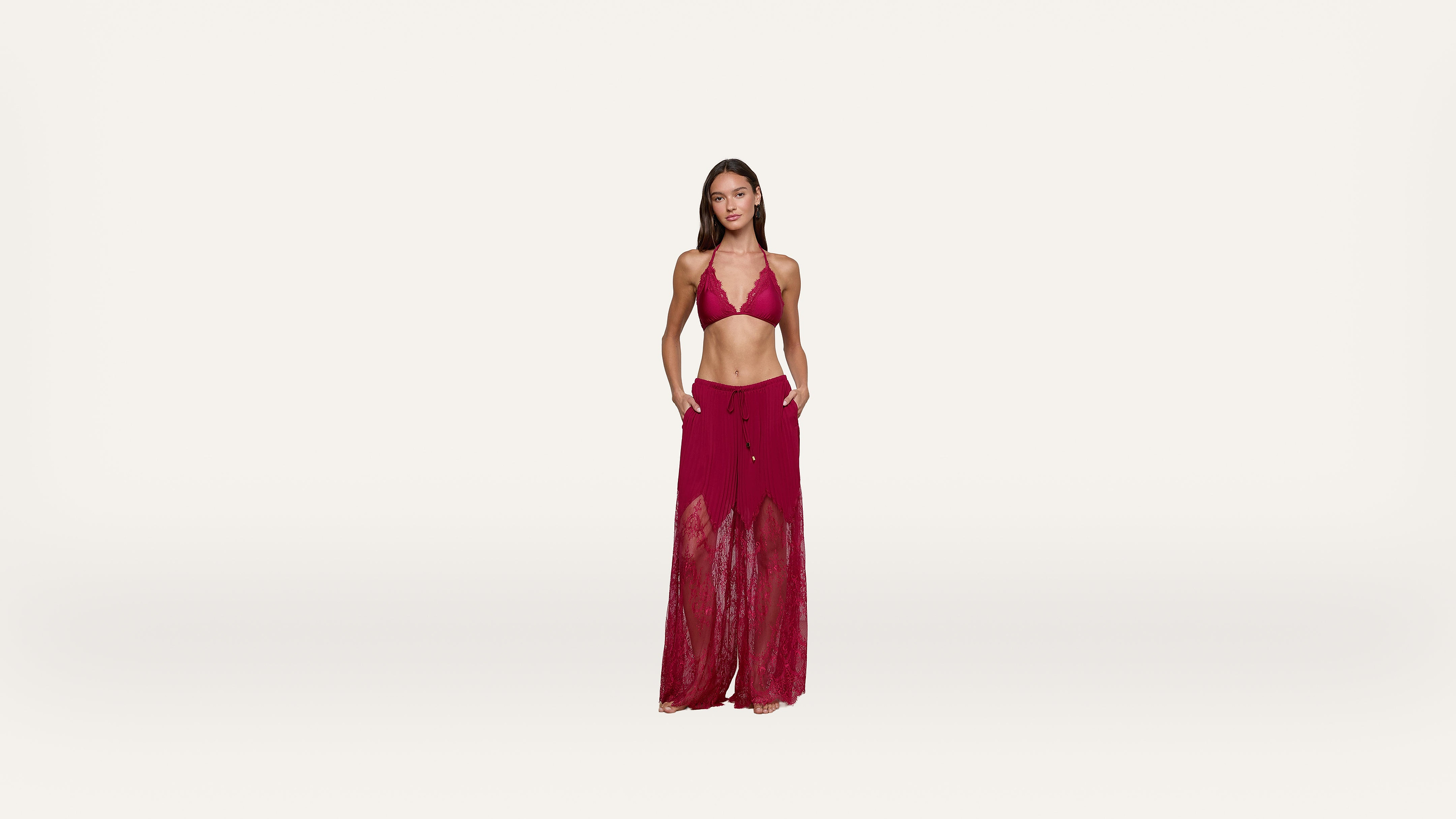 Woman standing against a plain white background, wearing a red halter bikini top and matching sheer, wide-leg beach pants with her hands in her pockets.