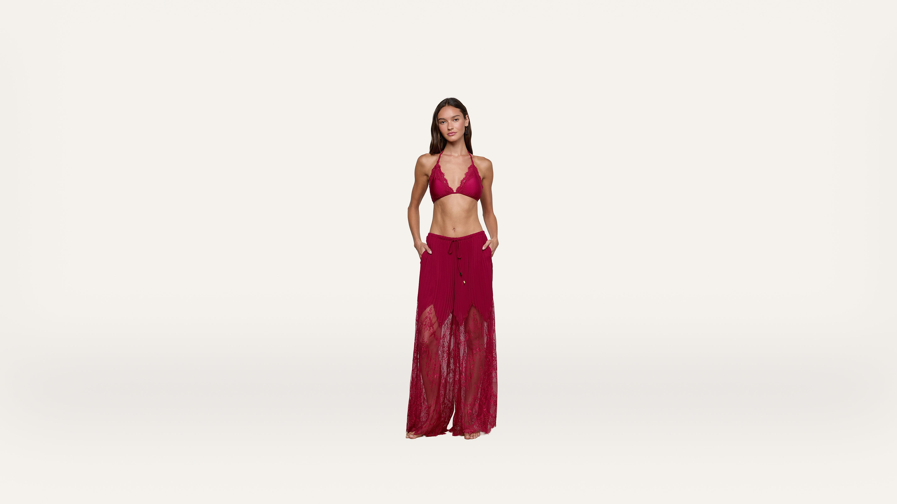 Woman standing against a plain white background, wearing a red halter bikini top and matching sheer, wide-leg beach pants with her hands in her pockets.