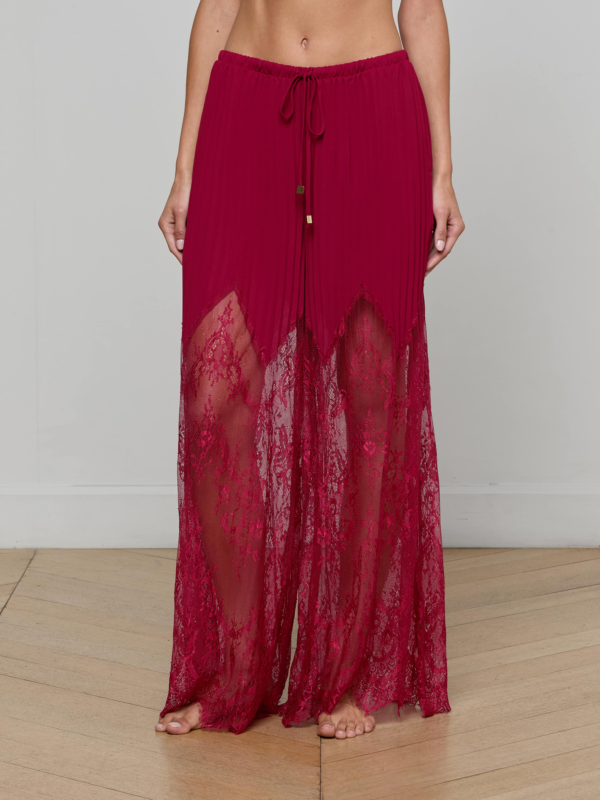 A person wearing the Sharon Cover-Up Pant—a long, sheer red lace pant with sheer lace panels and a drawstring waist, featuring solid fabric lining at the top—standing barefoot against a plain background.
