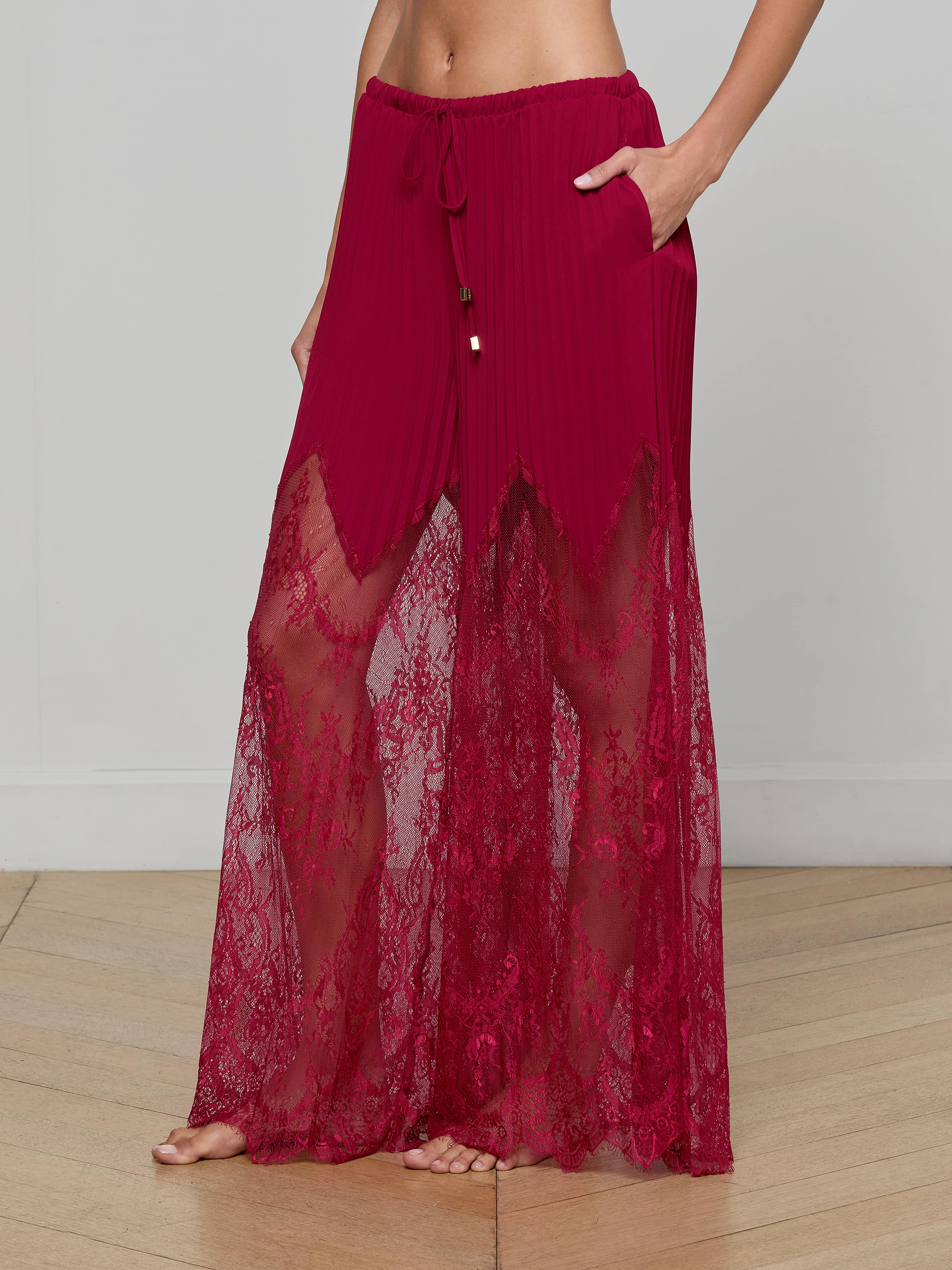 A person stands barefoot against a plain light background wearing the Sharon Cover-Up Pant, featuring sheer burgundy lace with a drawstring waist, pockets, and delicate sheer lace panels.