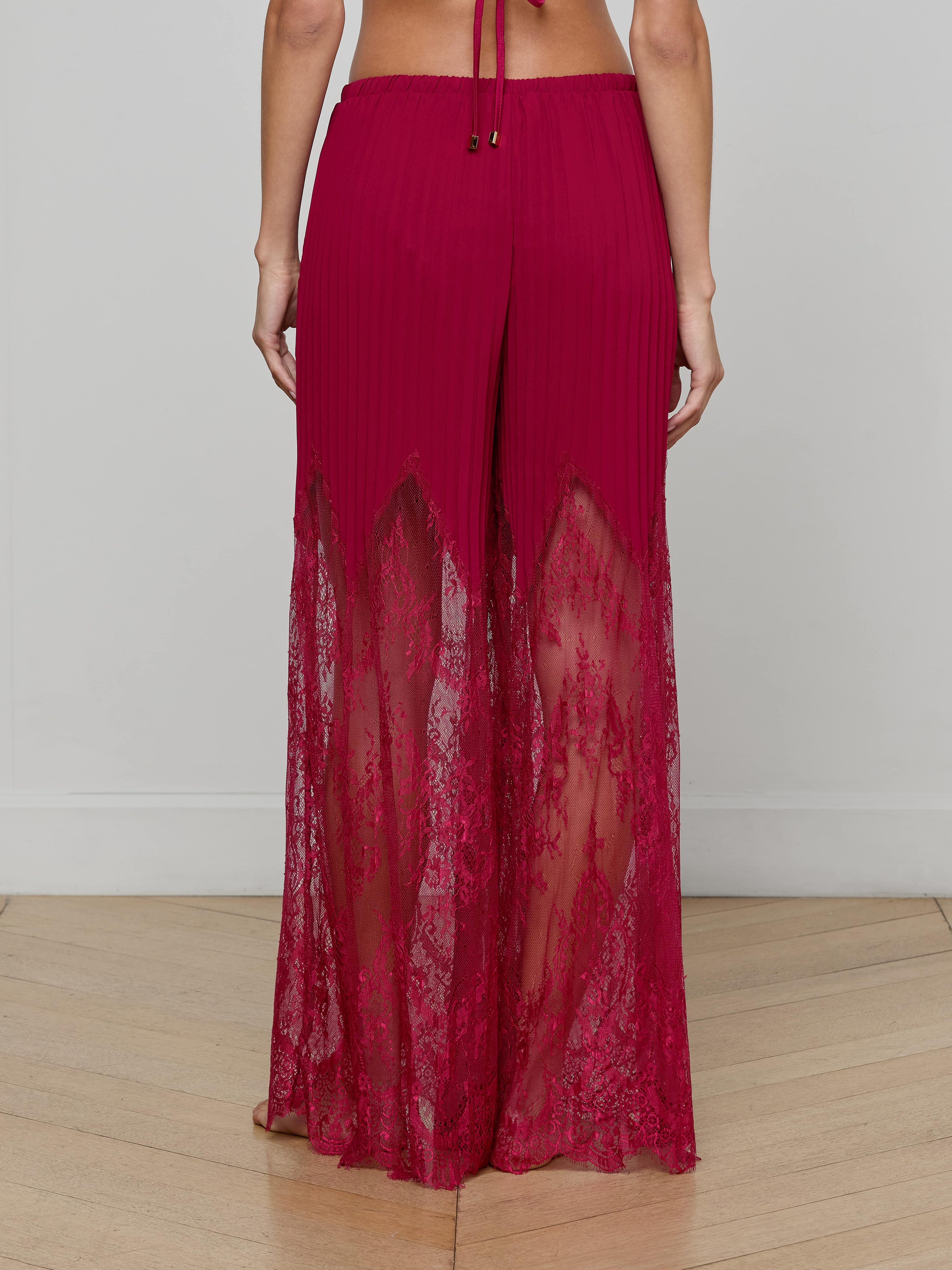 A person is shown from the back against a plain light background, wearing the Sharon Cover-Up Pant—high-waisted, wide-leg pants in red lace featuring sheer lace panels, a solid waistband, and a pleated upper section.