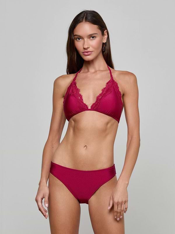 A woman stands against a plain background, wearing the Annabelle Triangle Bikini Top in deep red, featuring adjustable ties and lace trim. She has long brown hair and gazes at the camera with a relaxed expression.