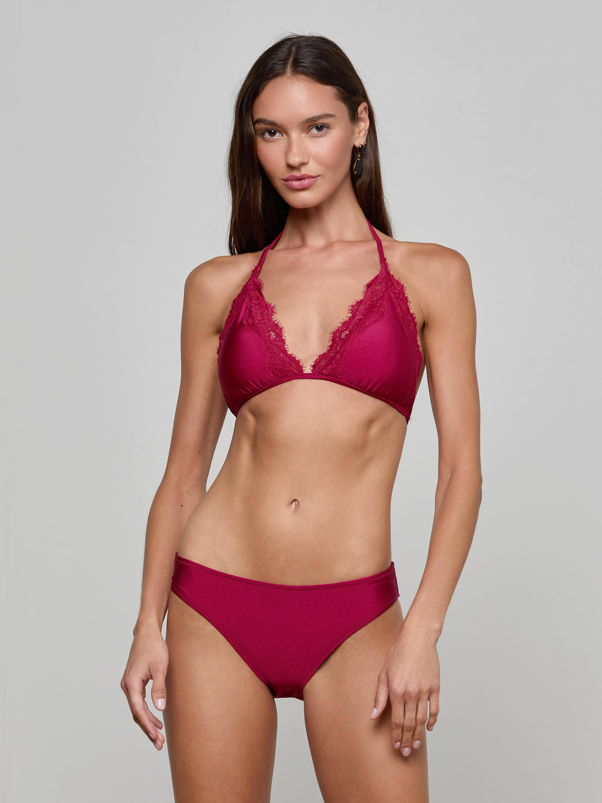 A woman stands against a plain background, wearing the Annabelle Triangle Bikini Top in deep red, featuring adjustable ties and lace trim. She has long brown hair and gazes at the camera with a relaxed expression.