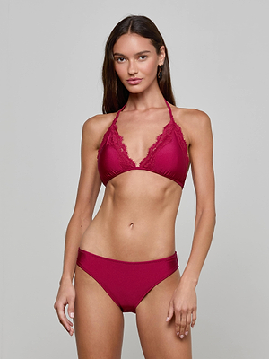 A woman with long brown hair models the Annabelle Triangle Bikini Top, featuring delicate lace trim and adjustable ties, while standing against a plain light gray background and looking at the camera.