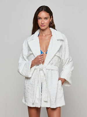 A woman with long brown hair wears the Caterina Cover-Up Robe, a textured white cotton jacquard belted coat, over a blue top, standing against a plain light gray background with one hand in her pocket.