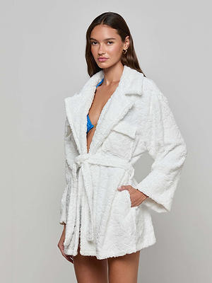 A woman wearing the Caterina Cover-Up Robe, a textured white cotton jacquard kimono, over a blue bikini poses with one hand in her pocket against a plain light gray background.