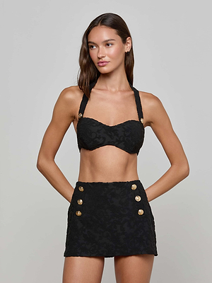 A woman stands with her hands on her hips, wearing the Coco Swim Skirt—a high-waisted mini with six gold buttons—paired with a black jacquard terry crop top with wide straps. The background is plain, light gray.