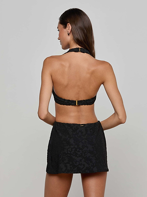 A woman faces away from the camera, wearing a black halter bikini top and the Coco Swim Skirt—a jacquard terry swim skirt with built-in bikini bottom. Her relaxed arms and long hair are visible against a plain light gray background.