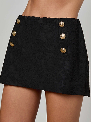 A close-up of a person wearing the Coco Swim Skirt, a black jacquard terry piece featuring gold decorative buttons in two vertical rows near the waist, with only their torso and upper legs visible.