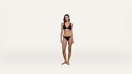 A woman with straight brown hair wears a black bikini and stands barefoot against a plain white background, looking directly at the camera with a neutral expression.