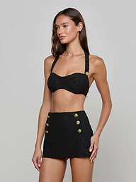 Wearing the Sally Bandeau Top and a matching black high-waisted mini skirt with gold buttons, a woman stands against a plain white background, exuding retro glamour.