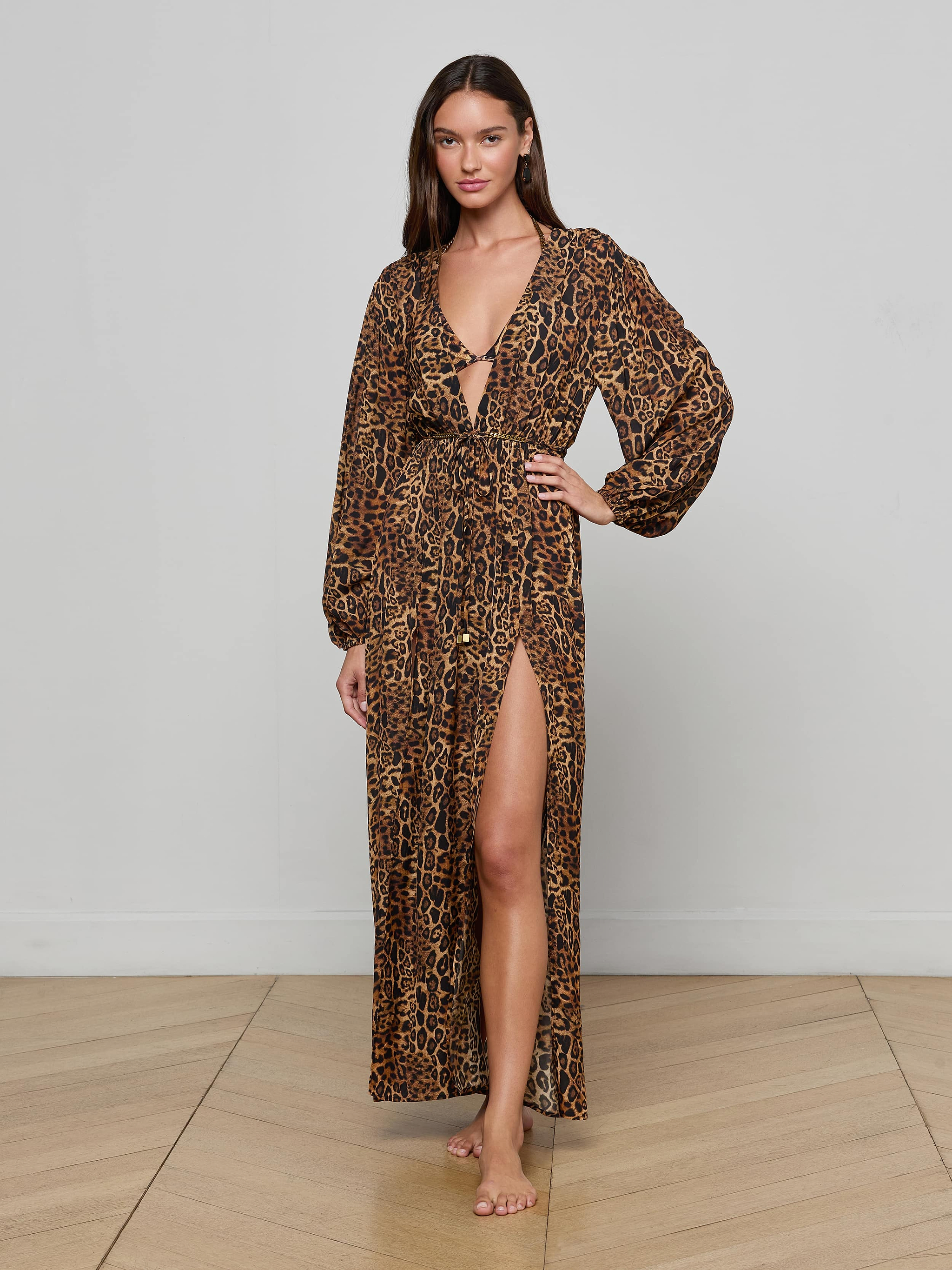 A woman stands barefoot against a plain white background, wearing the Sara Maxi Cover-Up Dress—a long-sleeved, leopard print piece featuring a deep V-neck and a high slit.