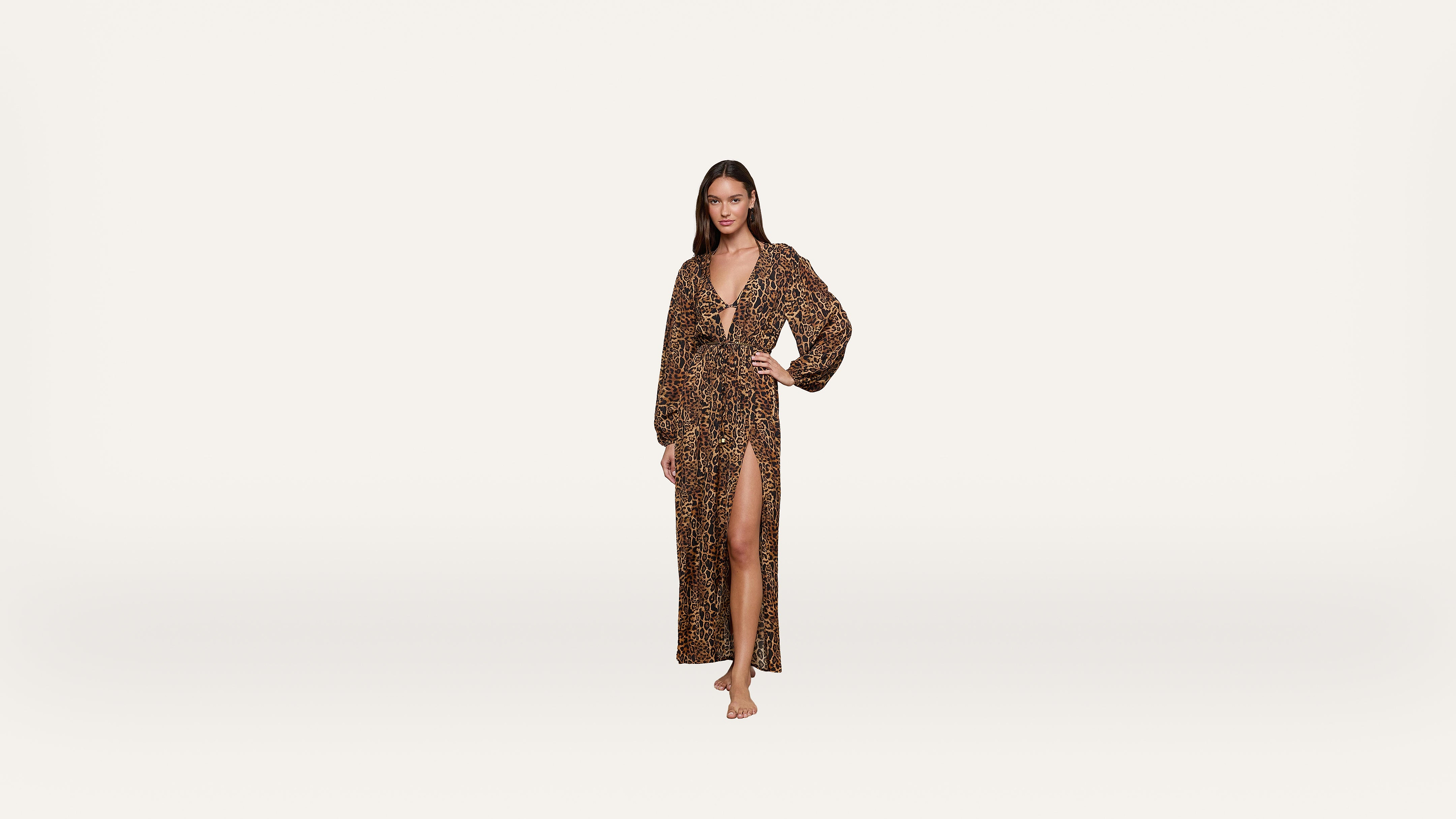 Woman stands barefoot wearing a long, leopard print dress with a deep V-neck, long sleeves, and a high slit up one leg, posing against a plain white background.