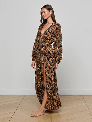 A woman stands barefoot on a wooden floor, wearing the L'AGENCE Sara Maxi Cover-Up Dress—a long, flowy leopard print cover-up with long sleeves, deep V-neckline, belted waist, and high slit—facing slightly left against a plain wall.