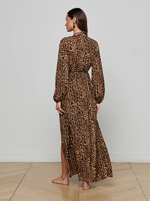A woman stands barefoot on a wooden floor, facing away from the camera, wearing the L'AGENCE Sara Maxi Cover-Up Dress—a flowy, long-sleeved leopard print piece—against a plain white wall.