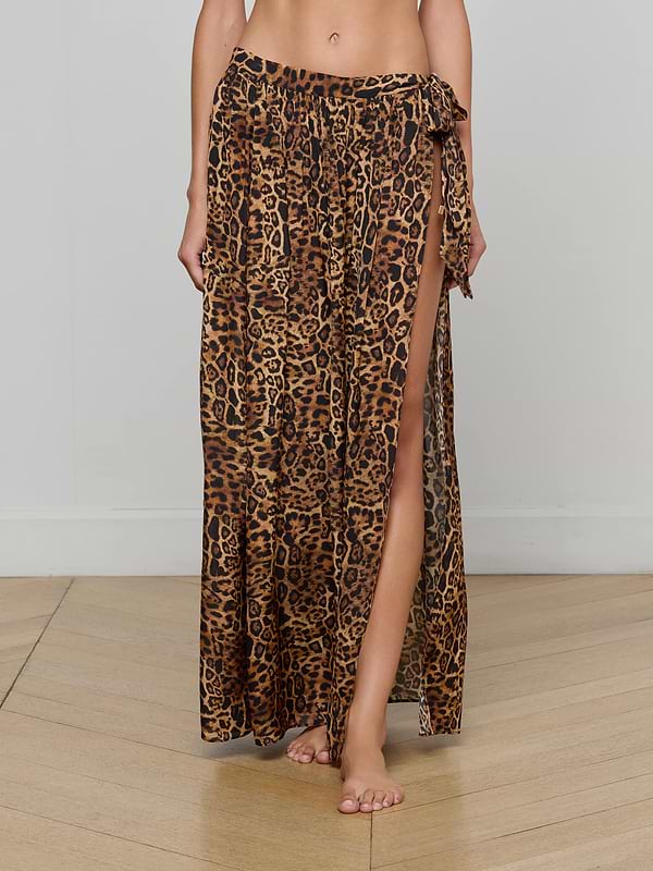A person wearing the Klaire Cover-Up Skirt—a long, high-slit wrap skirt in leopard print—stands barefoot against a plain background. Made from airy viscose chiffon, the skirt features a relaxed fit and ties at the waist.