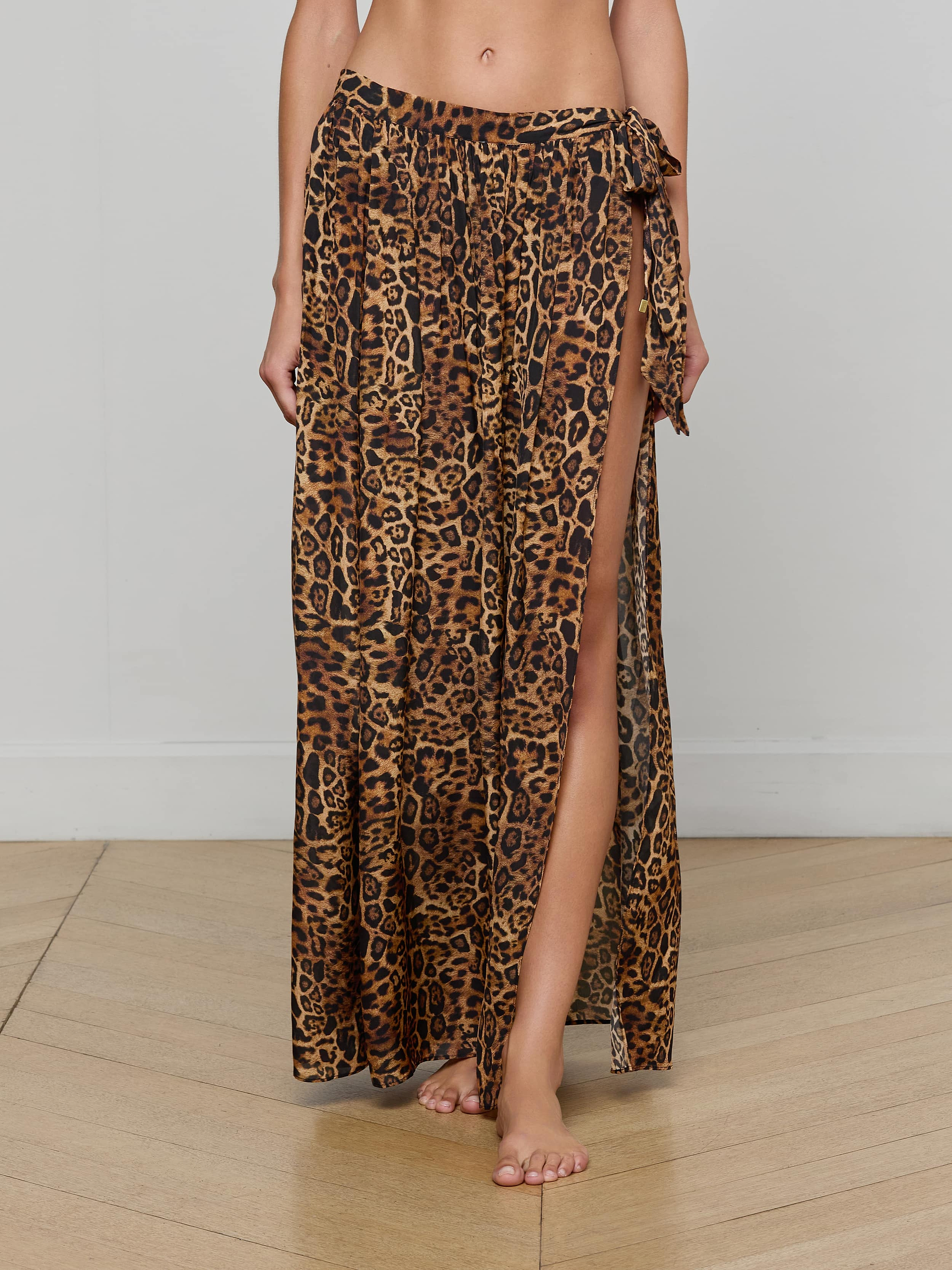 A person wearing the Klaire Cover-Up Skirt—a long, high-slit wrap skirt in leopard print—stands barefoot against a plain background. Made from airy viscose chiffon, the skirt features a relaxed fit and ties at the waist.