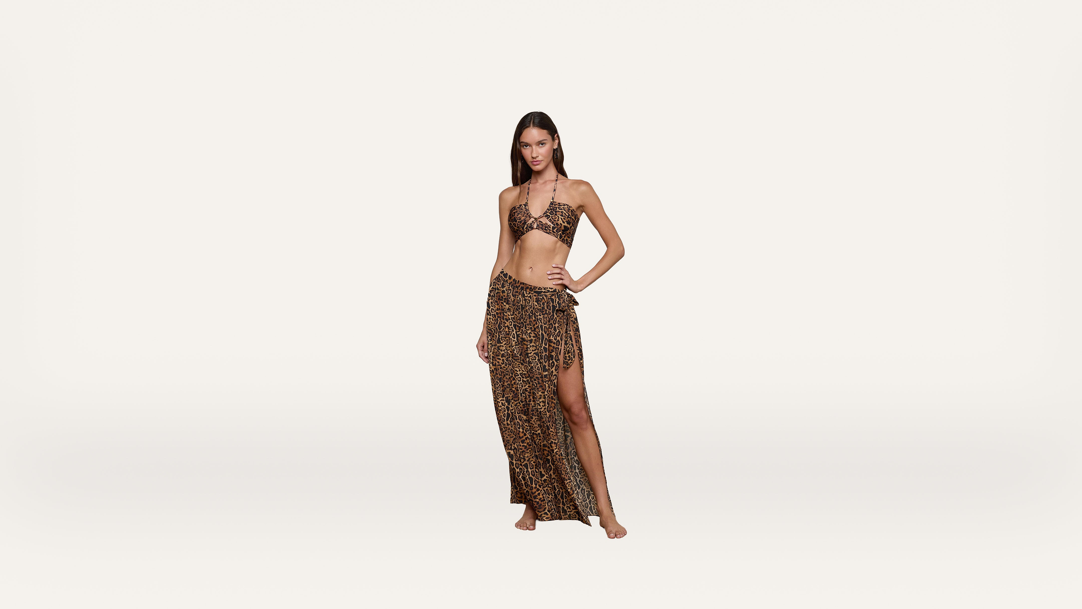 Woman wearing a leopard-print bikini top and matching high-slit sarong skirt, standing barefoot against a plain white background.